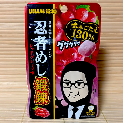 A black, red, and yellow 24g pouch of Ninja Meshi Tanren Red Grape gummies showing a glasses-wearing ninja mascot on a bamboo mat background.