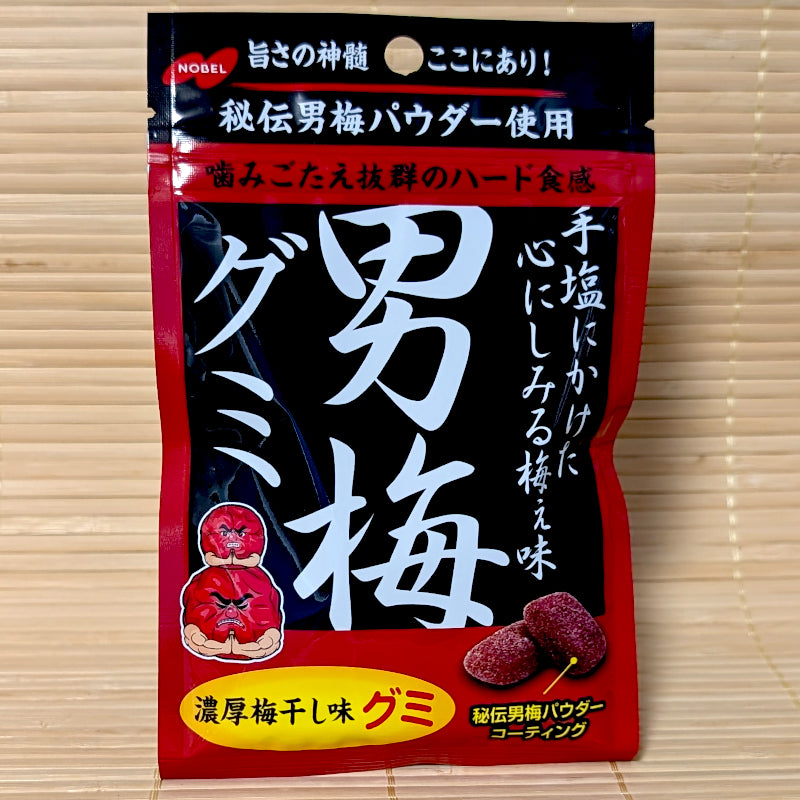 A bold black and red pouch of Nobel Otoko Ume Gummy candy featuring prominent Japanese calligraphy and the muscular red plum mascot, on a tatami mat background.