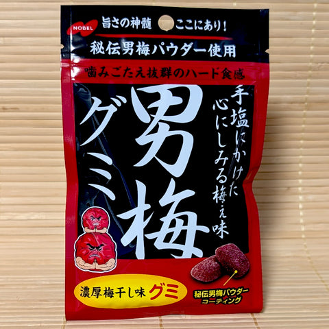 A bold black and red pouch of Nobel Otoko Ume Gummy candy featuring prominent Japanese calligraphy and the muscular red plum mascot, on a tatami mat background.