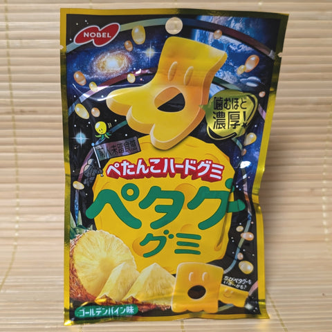 A metallic black and vibrant gold space themed pouch of Nobel Petagu Golden Pineapple gummy candy showing a flat yellow alien graphic on a tatami background.