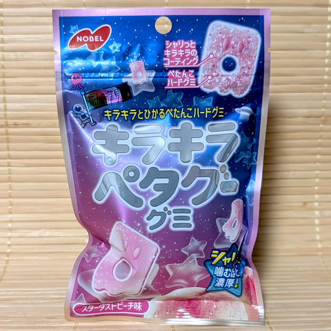A metallic purple and blue 50g pouch of Nobel Petagu Kira Kira Stardust Peach gummies showing an astronaut illustration, star graphics, and a sparkling pink flat gummy on a bamboo mat background.