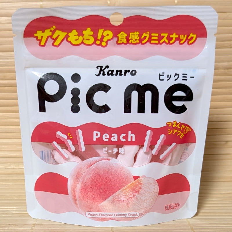 A white and pink pouch of Kanro Pic Me Peach gummy showing wavy dual-colored sticks and a large fresh peach graphic on a tatami background.