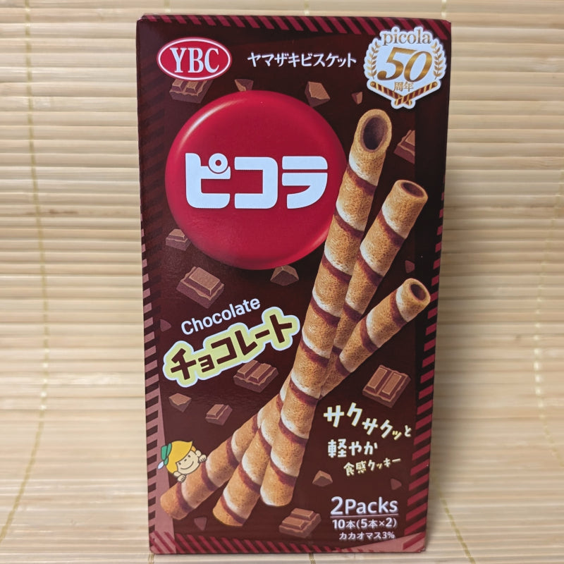 A box of Picola Cookie Sticks in milk chocolate flavor. The packaging is predominantly brown with an image of the cookie sticks and chocolate pieces. Displayed on a tatami background.

