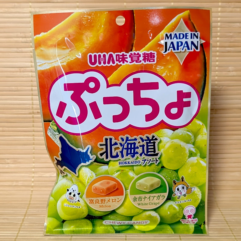 A metallic pouch of UHA Puccho Hokkaido Assortment showing orange Furano melon and green Niagara white grapes on a bamboo mat.