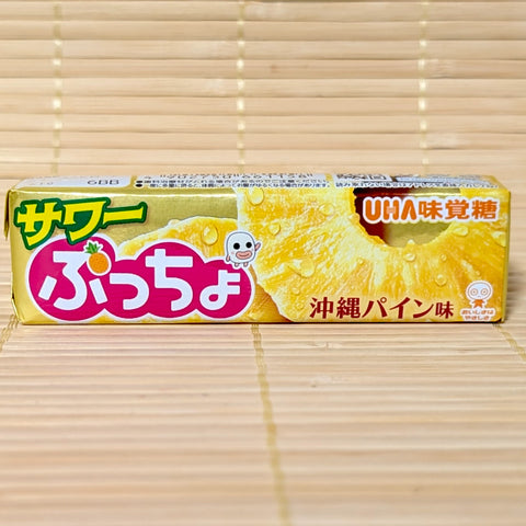 A metallic gold stick of UHA Sour Puccho Okinawa Pineapple candy showing a juicy cut pineapple graphic on a tatami background.