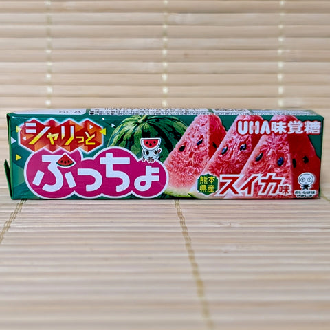 A green stick pack of UHA Puccho Suika Watermelon candy showing sliced red watermelons and a small mascot against a textured tatami background.