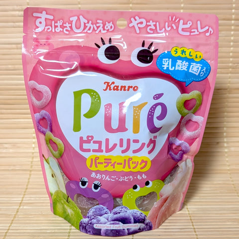 A bright pink 156g party pack bag of Kanro Puré Ring fruit gummies featuring cartoon character faces, heart-ring gummies, green apples, grapes, and peaches on a bamboo mat background.