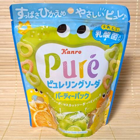A bright blue and green 156g party pack bag of Kanro Puré Ring Fruit Soda gummies showing cartoon character faces, heart-ring gummies, orange slices, muscat grapes, and pineapple on a bamboo mat background.