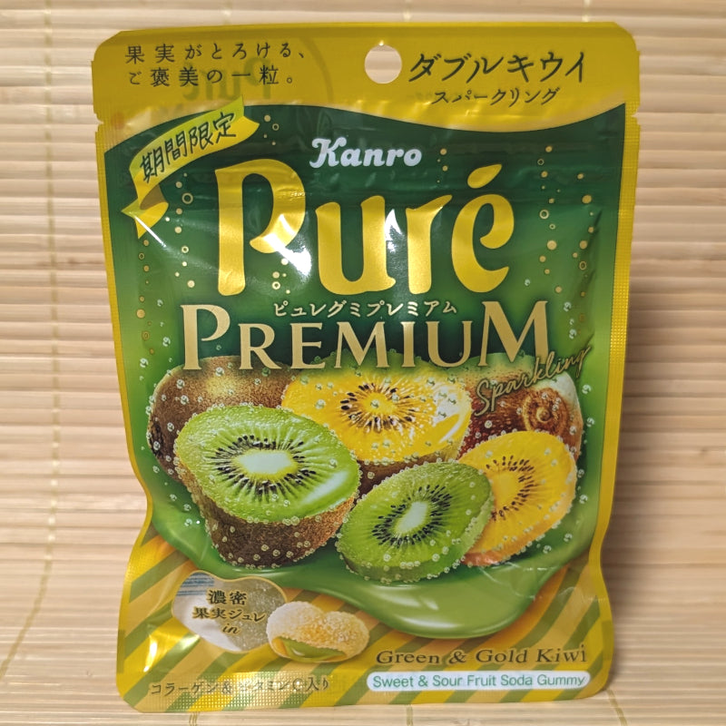 A metallic green and gold 54g pouch of Kanro Puré Premium Double Kiwi Sparkling gummies showing sliced green and gold kiwis, soda bubbles, and a cross-section of a heart-shaped gummy on a bamboo mat background.