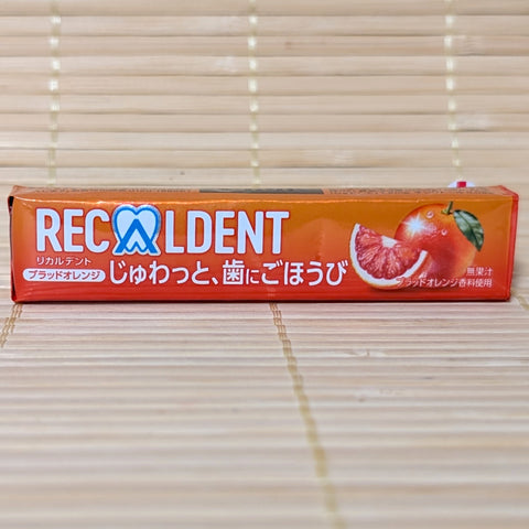 A metallic orange stick-pack of Recaldent Blood Orange sugarless gum displaying a sliced blood orange graphic on a bamboo mat background.
