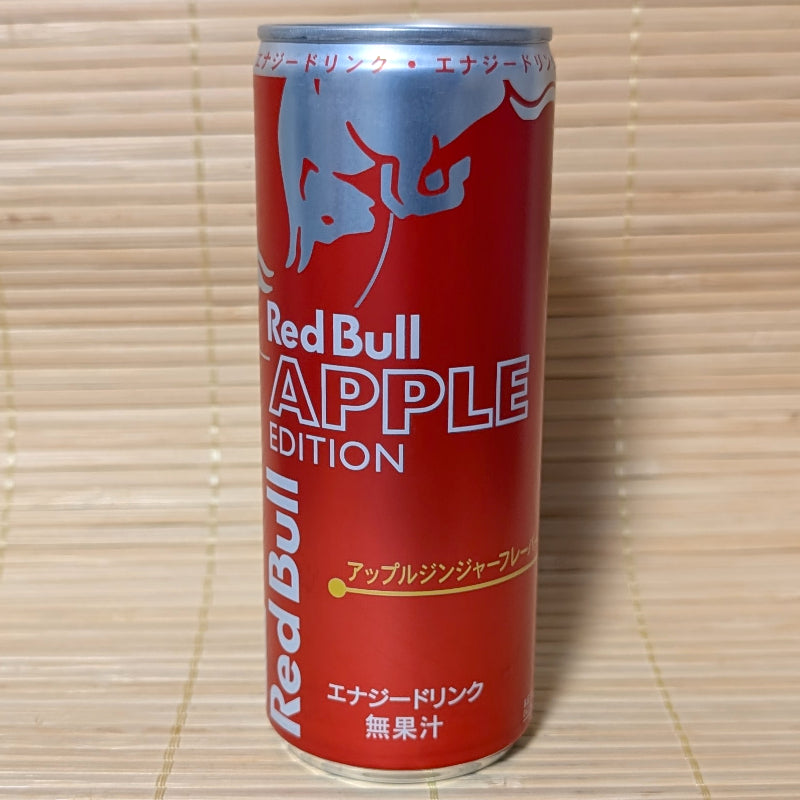 A solid red 250ml aluminum can of Japanese Red Bull Apple Edition showing silver bulls and gold katakana text on a bamboo background.   