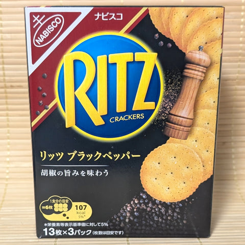 A black and gold Ritz box on a tatami background featuring a wooden pepper grinder, scattered peppercorns, and stacked Ritz crackers.