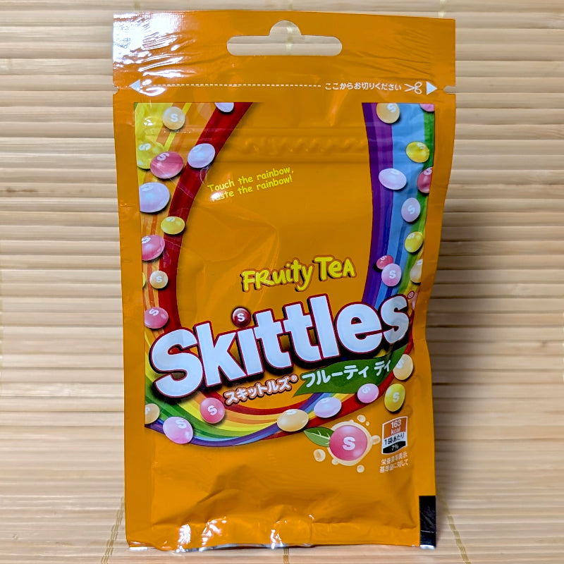 An orange resealable Skittles pouch on a tatami background featuring tea-themed "S" candies and a rainbow of fruity tea flavors.