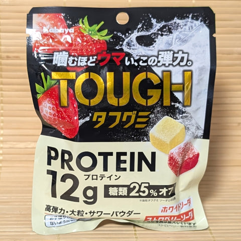 A black and cream 80g pouch of TOUGH Gummy Protein showing fresh strawberries, soda graphics, and cubed gummies on a bamboo background.   