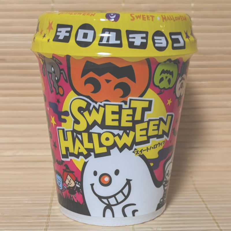 A bright yellow and white Halloween foam cup filled with 34 Tirol chocolates showing ghosts and pumpkins on a bamboo mat background.