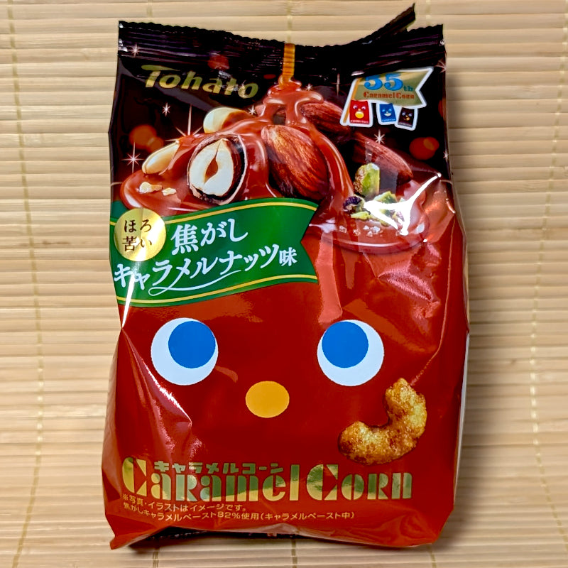 A dark red and black 55g bag of Tohato Caramel Corn Burnt Caramel Nuts flavor with almonds and hazelnut graphics on a bamboo mat.