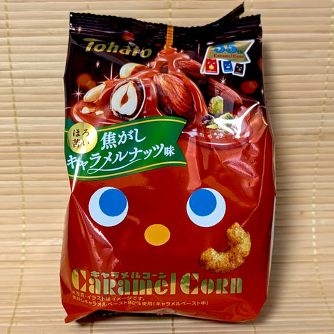 A dark red and black 55g bag of Tohato Caramel Corn Burnt Caramel Nuts flavor with almonds and hazelnut graphics on a bamboo mat.