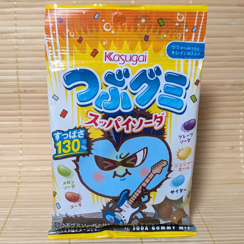 A yellow and blue 70g pouch of Kasugai Tsubugumi Suppai Soda Gummy Mix showing a cool blue mascot playing an electric guitar, a 130% sourness badge, and individual gummy beans representing five soda flavors on a bamboo mat background.
