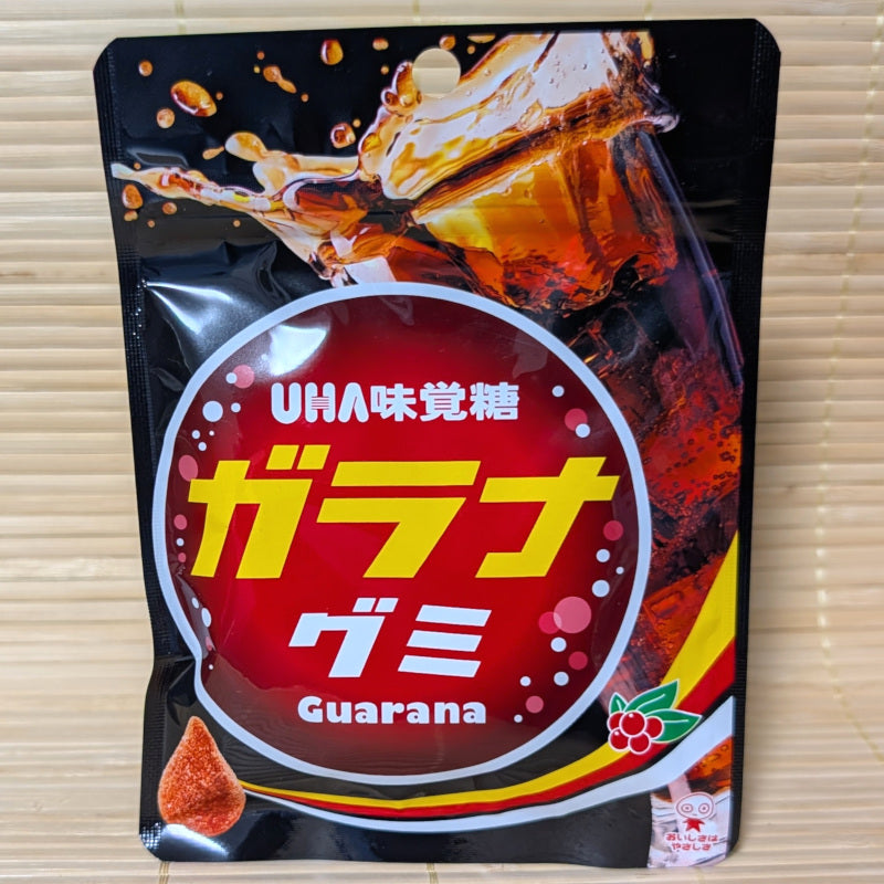 A black and red bag of UHA Guarana gummies featuring a splash of soda and a single teardrop shaped gummy with sugar coating.