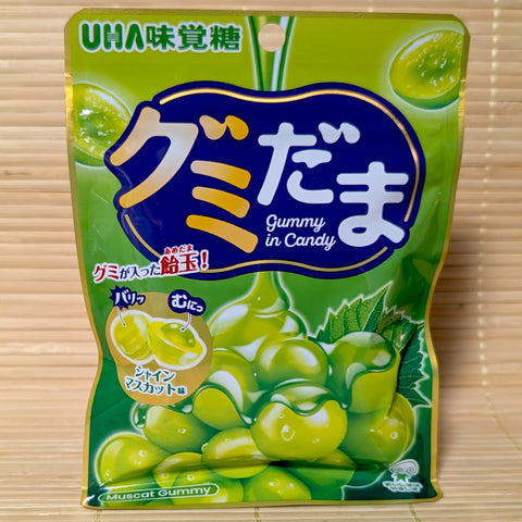 A green and gold 55g resealable pouch of UHA Gumidama Shine Muscat grape candy showing crunchy shell cross-section art on a bamboo mat background.
