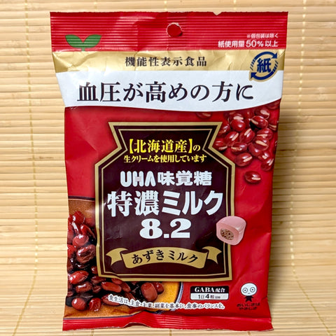 A red and gold 93g paper bag of UHA Tokuno Milk 8.2 Azuki Milk showing red beans, Hokkaido cream, and a cross-section of the candy on a bamboo mat background.