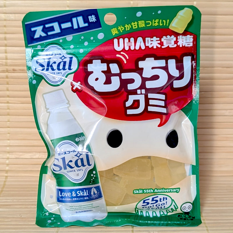 A green and red 64g pouch of UHA Mikakuto Mucchiri Gummy Skål Soda showing a Skål bottle illustration, a 55th Anniversary logo, and bottle-shaped gummies on a bamboo mat background.
