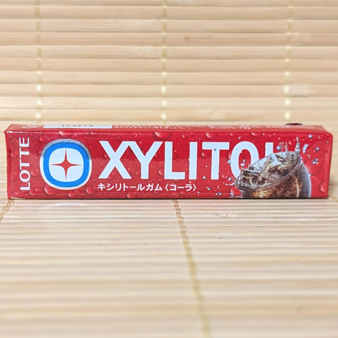 A red 14-piece stick pack of Lotte Xylitol Cola chewing gum featuring ice-cold soda graphics on a bamboo mat background.