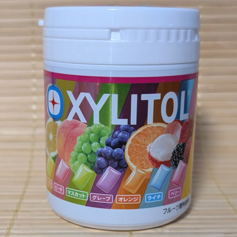 A large white plastic bulk tub of Lotte Xylitol gum showing a rainbow background with fresh peach, muscat, grape, orange, lychee, and berry graphics on a tatami background.